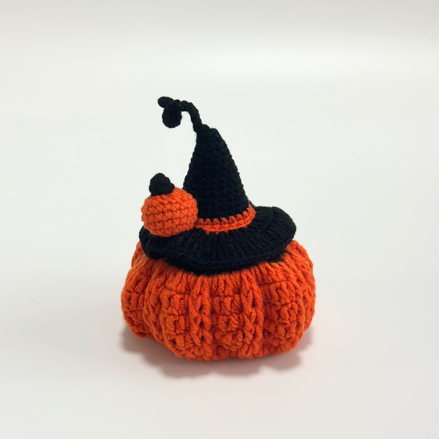 Side view of handmade crocheted Halloween pumpkin costume for Labubu doll showing curved black witch hat with orange pom-pom and trim detail, orange textured pumpkin body with ribbed crochet pattern. Empty costume displays construction quality and dimensional shape, perfect collectible Halloween doll accessory for craft enthusiasts and toy collectors