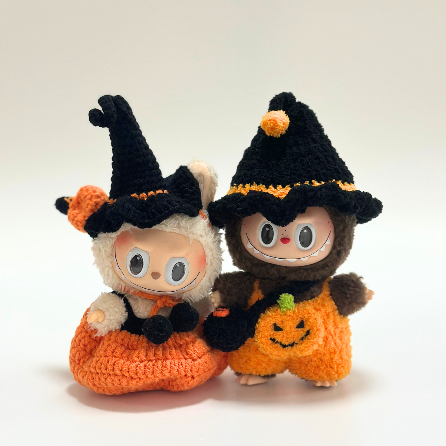 Close-up of two Labubu dolls wearing handmade crocheted Halloween witch costumes with black pointed hats featuring orange pom-poms and trim, orange pumpkin jumpsuits with embroidered jack-o'-lantern faces and green stem details, plus black accessories. Adorable Halloween doll duo, matching seasonal outfits, collectible miniature costumes perfect for Halloween enthusiasts and doll collectors.