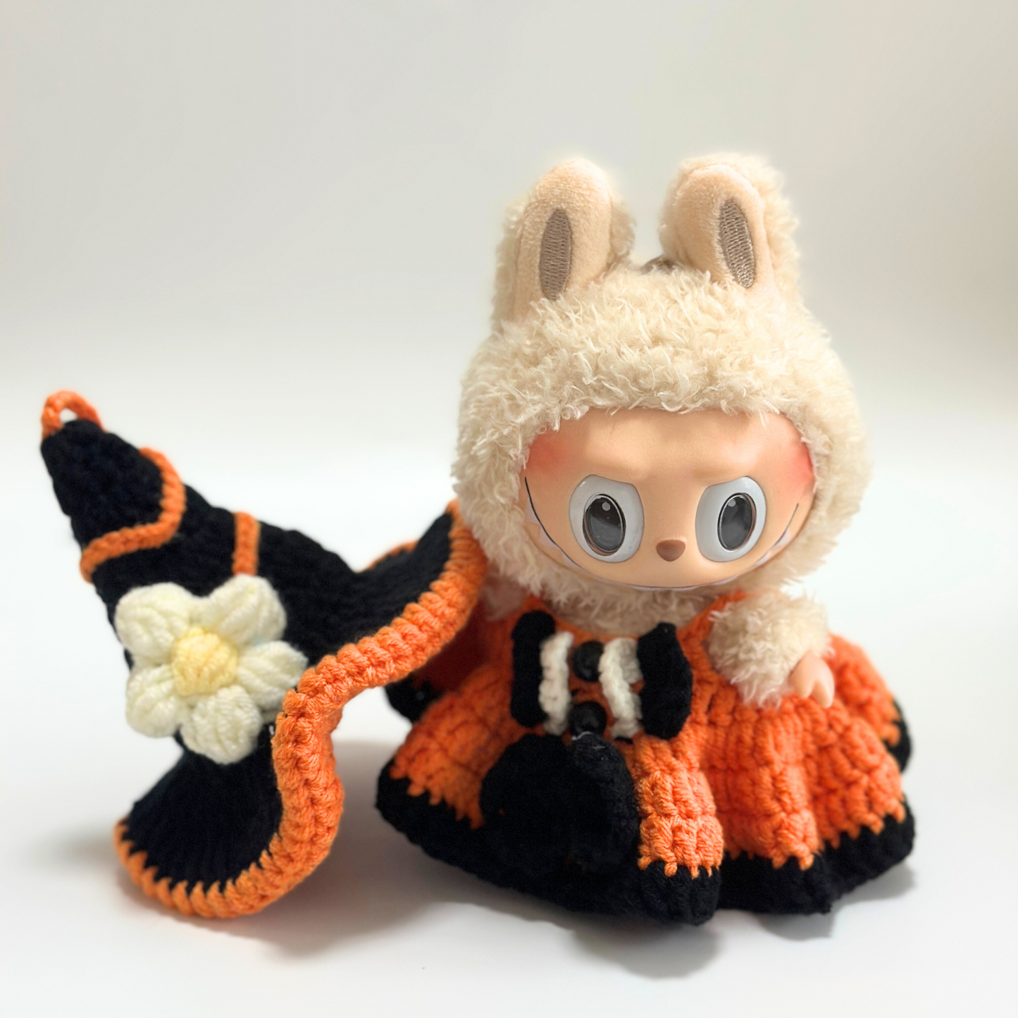 Labubu doll in handmade crocheted Halloween witch costume holding black witch hat with orange trim and white daisy flower decoration. Doll wears orange textured dress with black trim details and matching accessories. Shows intricate flower embellishment and quality craftsmanship of Halloween doll costume, perfect collectible seasonal outfit for toy enthusiasts and craft lovers.