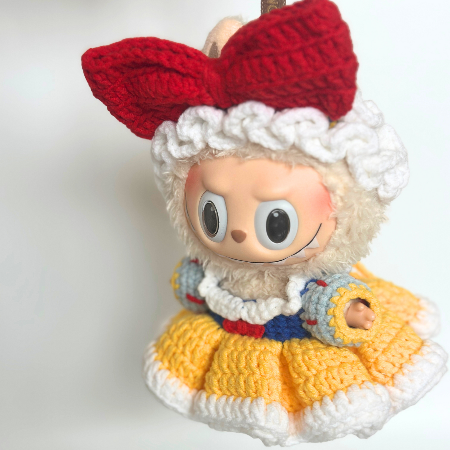 Close-up detail view of a crocheted Snow White costume on a Labubu doll, showing the intricate stitching of the red bow headband, white ruffled bonnet trim, yellow textured skirt with detailed crochet patterns, blue bodice with red accents, and white puffy sleeves. The doll's characteristic large round eyes and gentle smile are prominently featured.