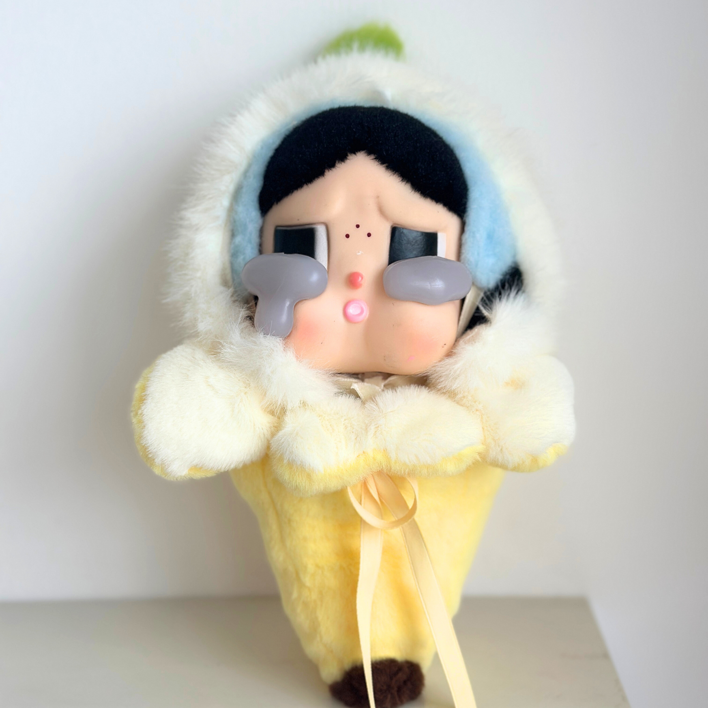 Crybaby doll dressed in adorable banana costume featuring soft white hood with green accent, yellow plush body