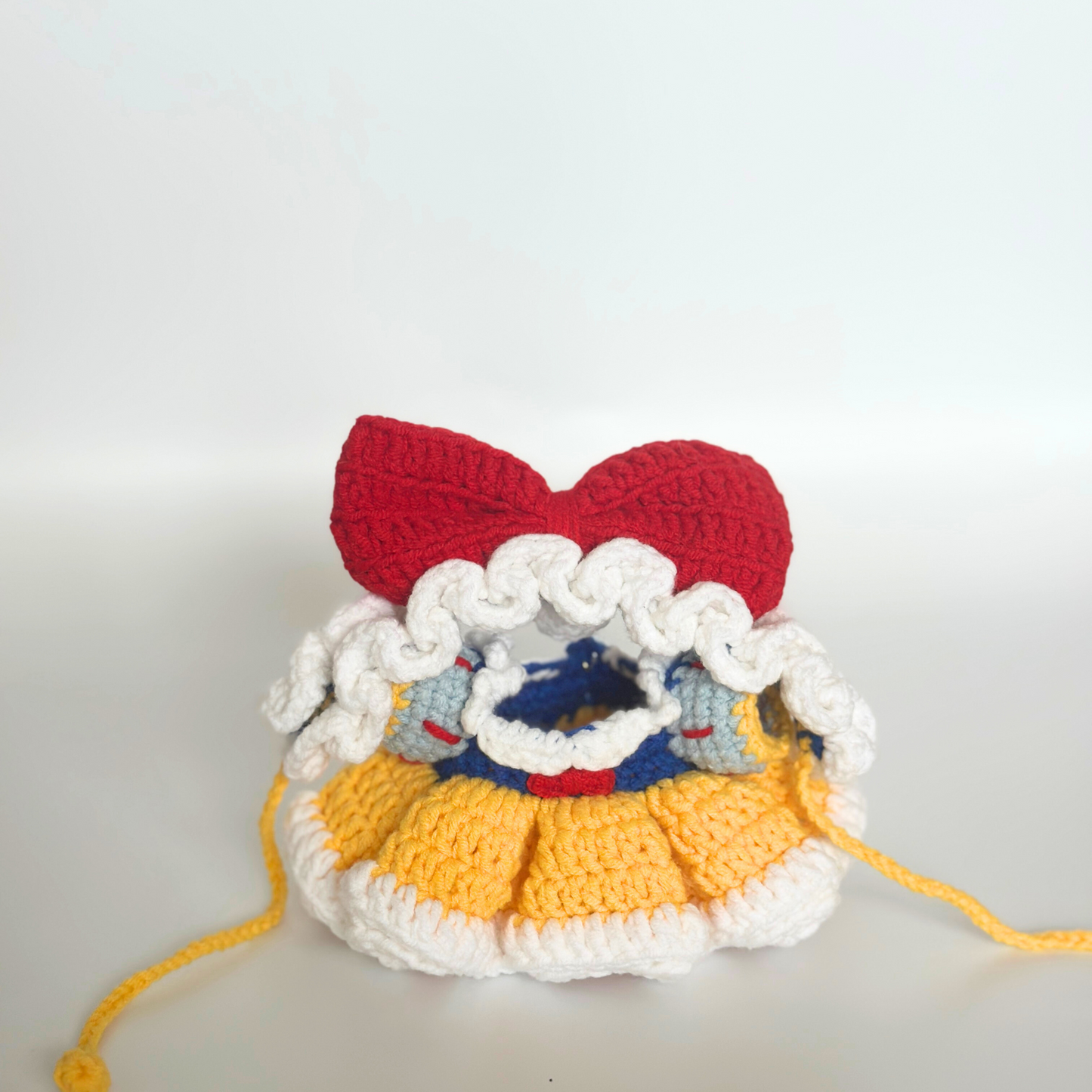 Front view of a crocheted Snow White costume for Labubu doll without the doll inside, showing the red bow headband at top, white ruffled bonnet trim, blue bodice with white collar and red accents, layered yellow ruffled skirt, white base, and yellow drawstring ties. The empty costume displays the intricate crochet construction and dimensional details of each component.