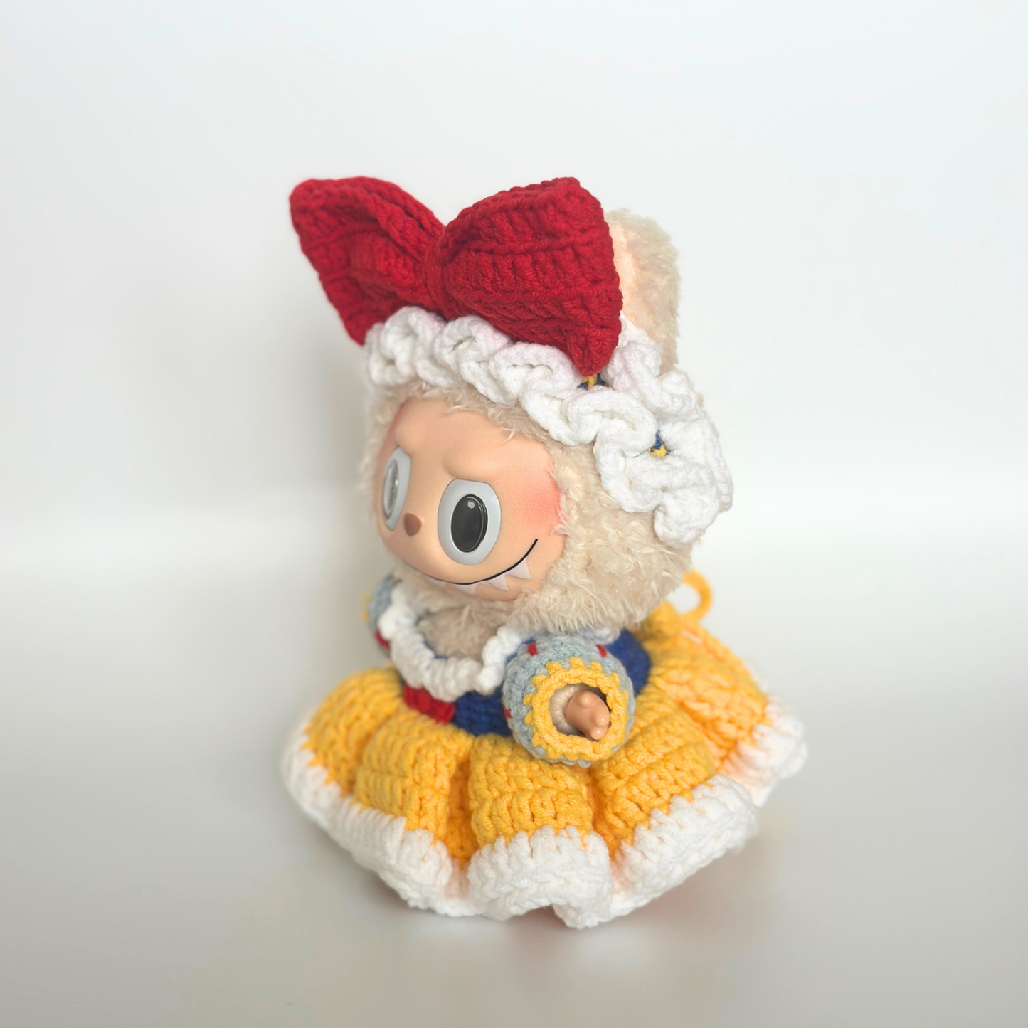 Side profile view of a Labubu doll wearing a handmade crocheted Snow White costume, showcasing the dimensional red bow headband, white ruffled bonnet with decorative trim, layered yellow and white ruffled skirt, and blue bodice with puffy white sleeves. The doll displays its characteristic sweet expression with large round eyes against a clean light background