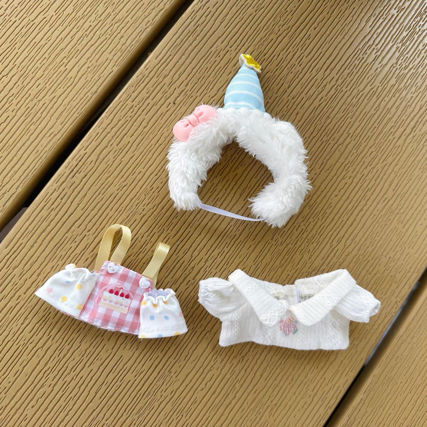 Three main components of Labubu birthday costume - white fluffy hood with striped party hat, checkered apron with cake design, and white ruffled off-shoulder top laid out on wooden deck surface