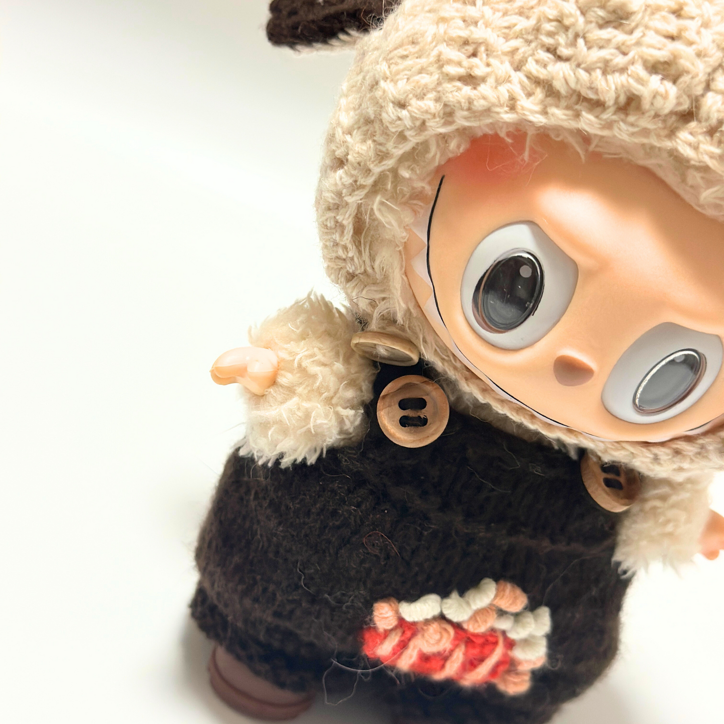 Close-up detail of Labubu doll in cream bunny ear hoodie and brown overalls showing wooden button details and embroidered popcorn design