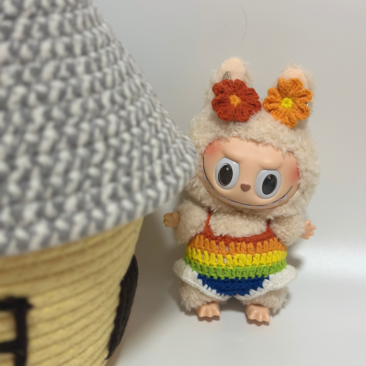 Labubu doll in handmade rainbow gradient knitted dress with orange crocheted flower headband, positioned next to woven wicker basket on white surface