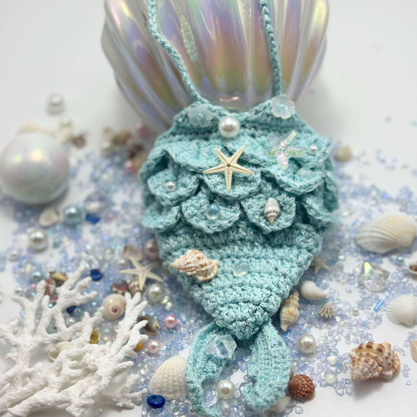 Labubu doll mermaid costume styled in ocean-themed scene with mint green crocheted scales decorated with starfish, pearls, and seashell accessories, surrounded by white coral, colorful seashells, pearls, and iridescent backdrop. Shows detailed craftsmanship with ocean-themed embellishments creating magical underwater fantasy display, perfect collectible mermaid costume for doll enthusiasts.