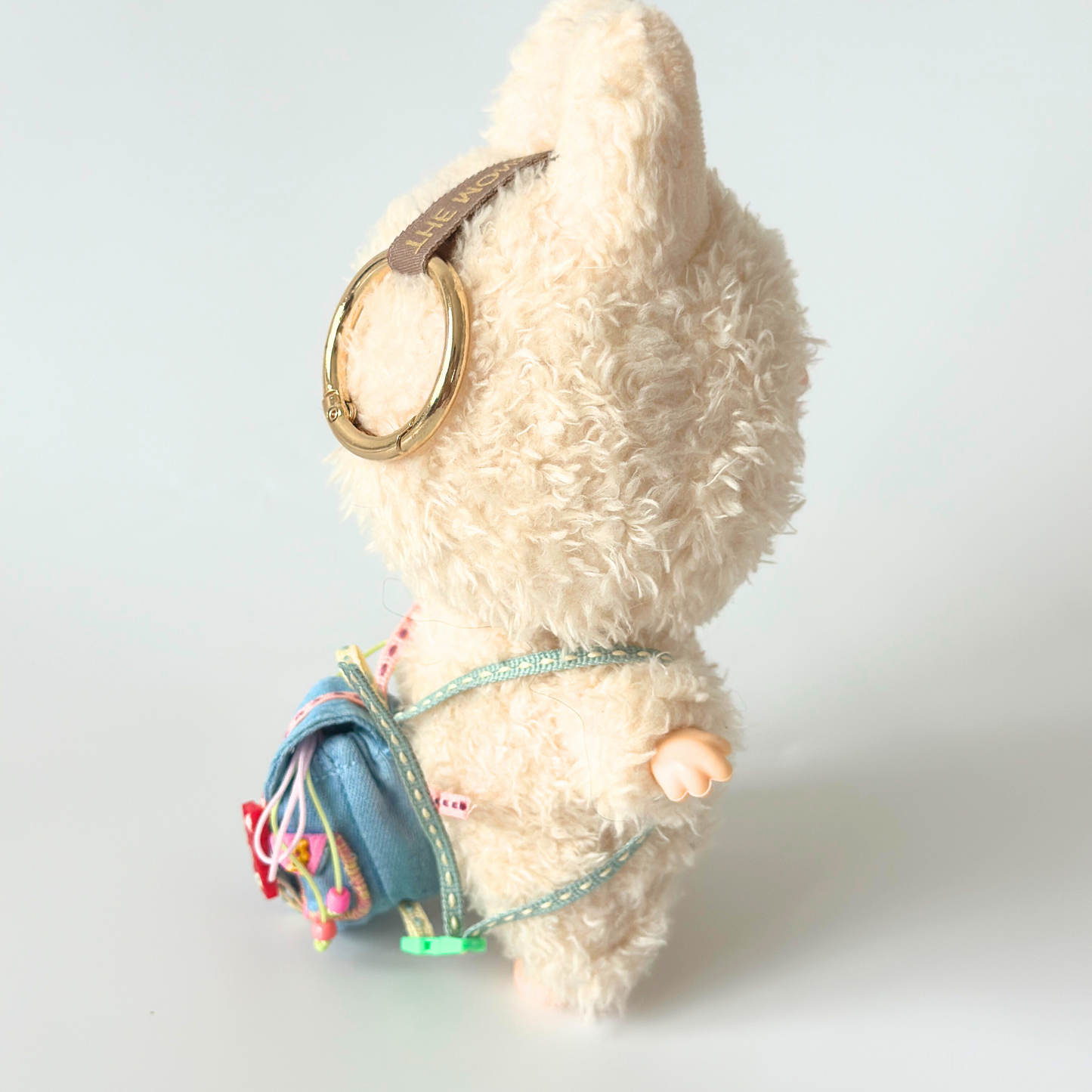 Side profile view of Labubu doll wearing blue corduroy miniature backpack with colorful rainbow straps and decorative ribbons, gold ring accessory on head with brown ribbon detail. Shows realistic proportions and quality craftsmanship of handmade doll accessories, clean white background highlighting the miniature backpack construction and functional straps.
