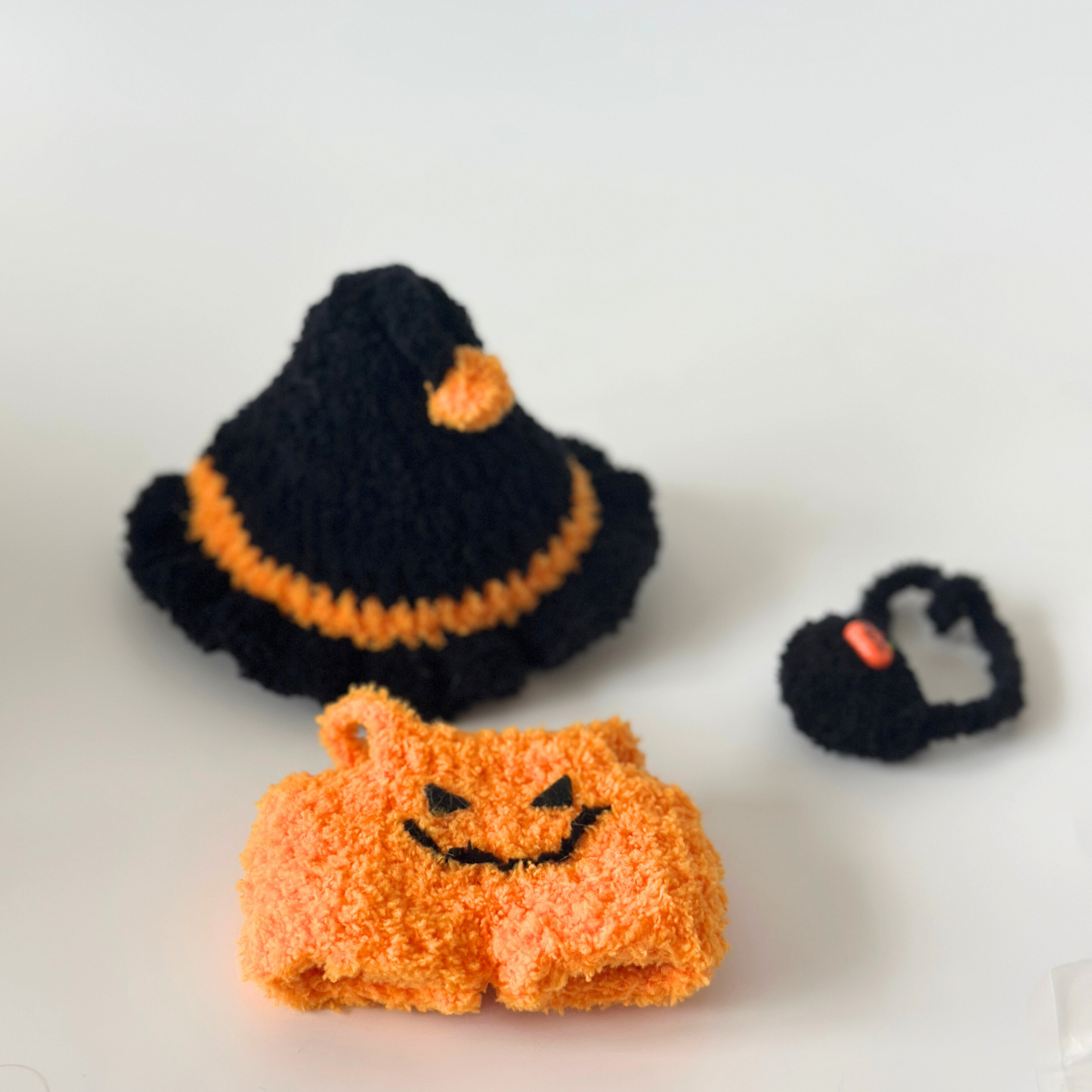 Flat lay of handmade crocheted Halloween costume components for Labubu doll including black witch hat with orange pom-pom and trim details, orange pumpkin jumpsuit with embroidered jack-o'-lantern smile face, and black trick-or-treat bag with pumpkin charm. Complete Halloween doll accessory set, collectible seasonal craft items for toy enthusiasts and doll collectors.