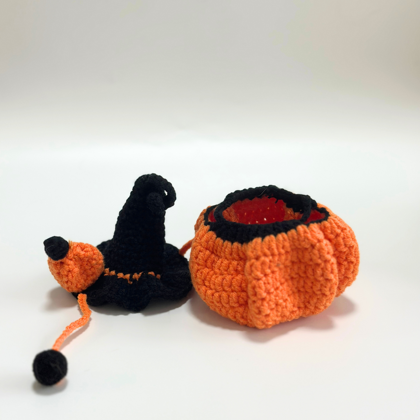 Open view of handmade crocheted Halloween pumpkin costume for Labubu doll showing interior red lining and construction details, with black witch hat featuring curved tip and orange pom-pom alongside. Orange textured pumpkin body displays quality craftsmanship and dimensional shape with drawstring closure, perfect collectible Halloween doll costume for craft enthusiasts.