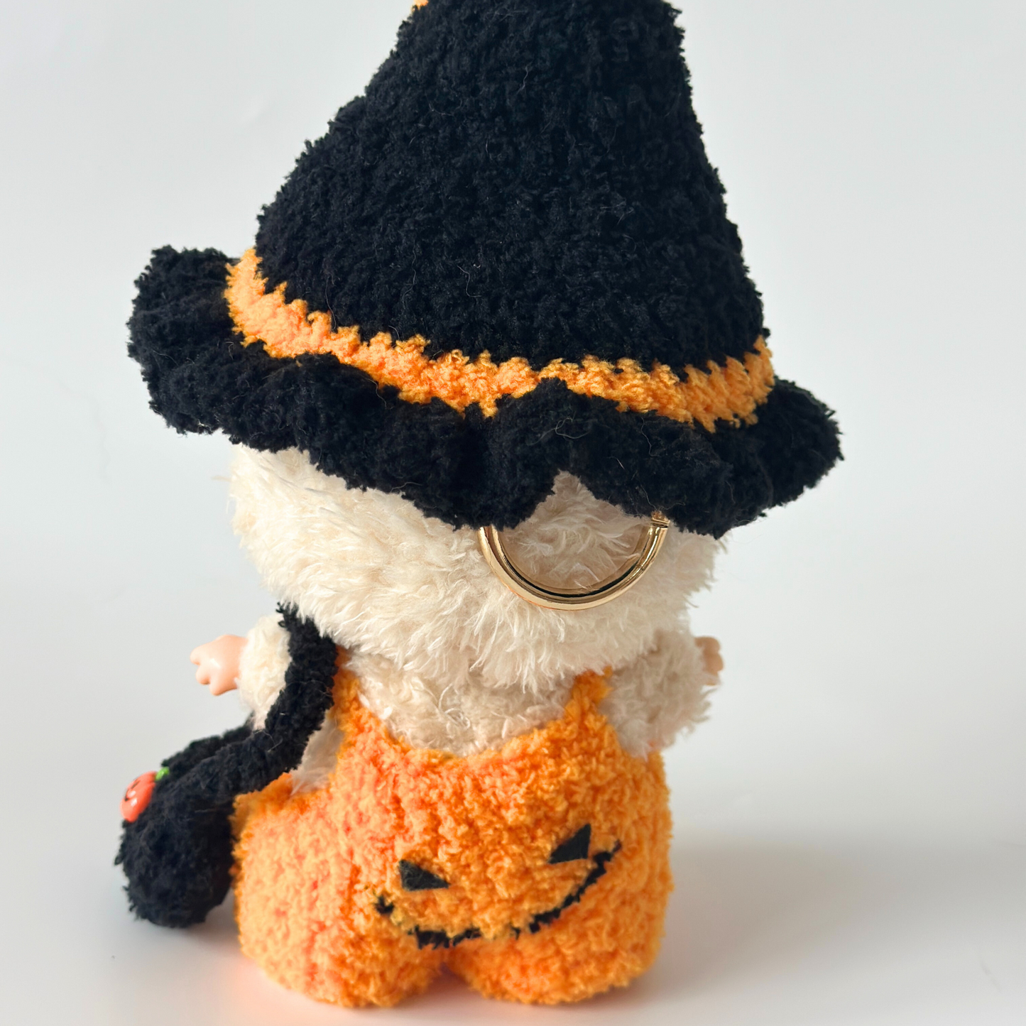 Back view of Labubu doll wearing handmade crocheted Halloween costume showing black witch hat with orange pom-pom and trim details, orange pumpkin jumpsuit with embroidered jack-o'-lantern face on back, black trick-or-treat bag with pumpkin charm, and gold ring detail. Perfect Halloween doll costume, collectible seasonal outfit for toy enthusiasts and craft lovers.