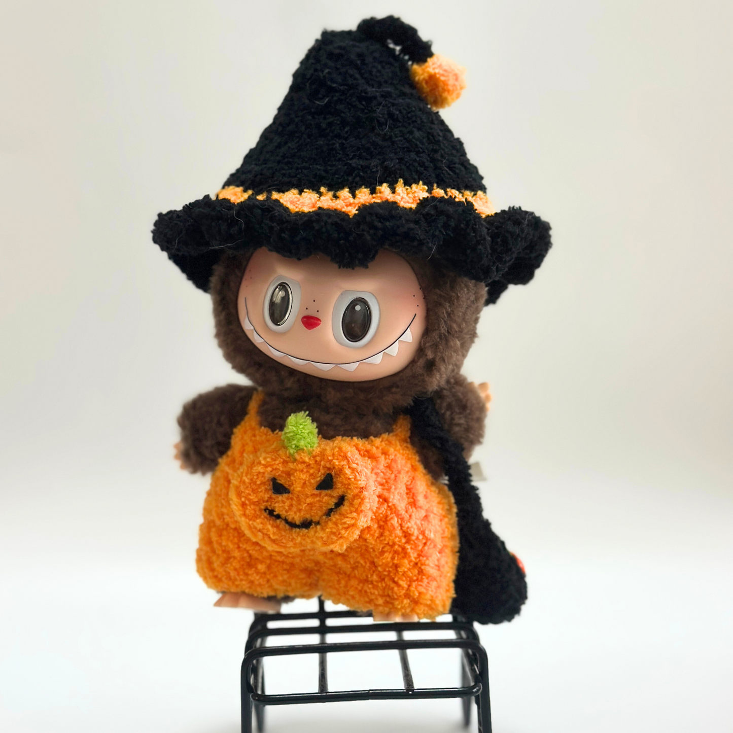 Labubu doll wearing handmade crocheted Halloween costume sitting on miniature black wire chair. Features black witch hat with orange pom-pom trim, orange pumpkin jumpsuit with embroidered jack-o'-lantern face and green stem detail, plus black trick-or-treat bag. Adorable Halloween doll photography, collectible seasonal outfit for toy collectors and craft enthusiasts.