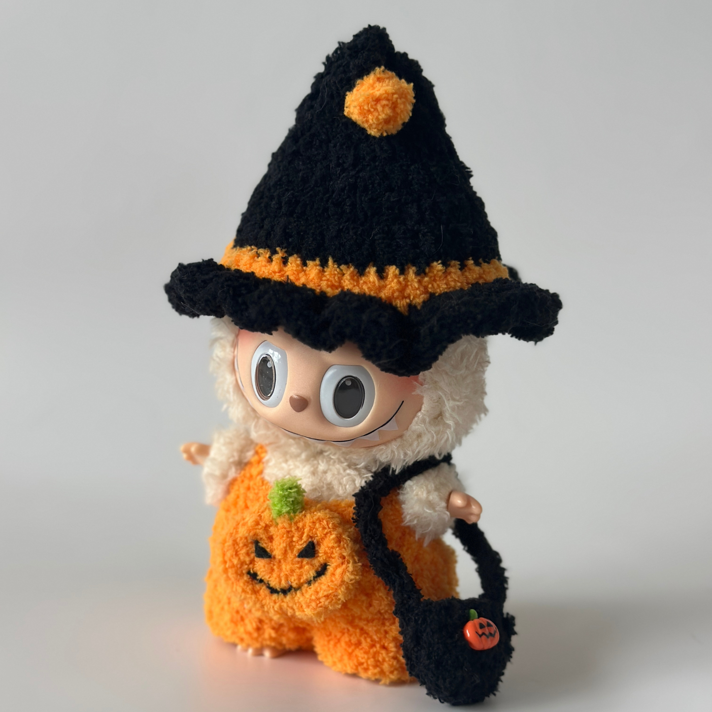 Handmade crocheted Halloween costume for Labubu doll featuring black witch hat with orange pom-pom and trim, orange pumpkin jumpsuit with embroidered jack-o'-lantern face and green stem detail, plus black trick-or-treat bag with pumpkin charm. Perfect Halloween doll outfit, collectible seasonal costume for toy enthusiasts.