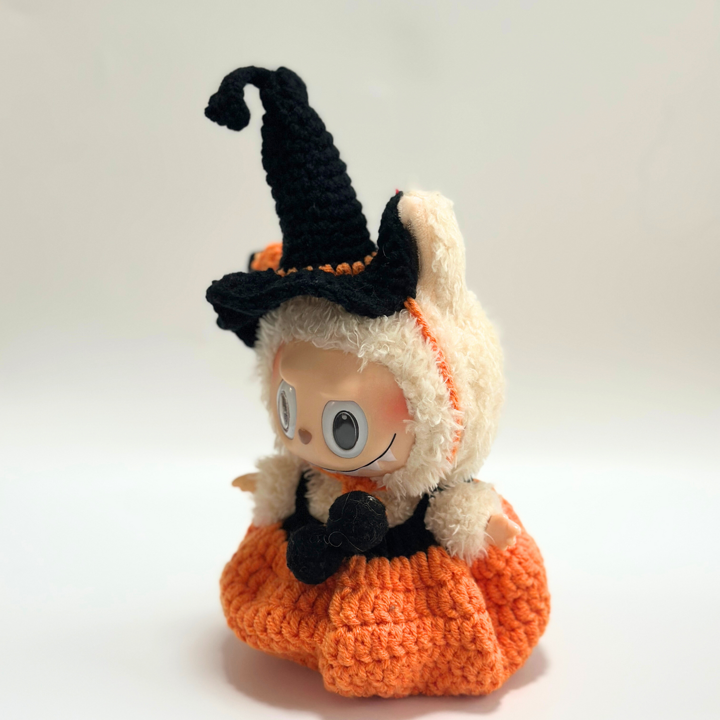 Side profile view of Labubu doll wearing handmade crocheted Halloween witch costume with curved black pointed hat featuring orange trim, orange textured pumpkin dress with ribbed crochet pattern, and black bow accessory. Shows intricate construction details and dimensional hat shape, perfect collectible Halloween doll outfit for toy enthusiasts.