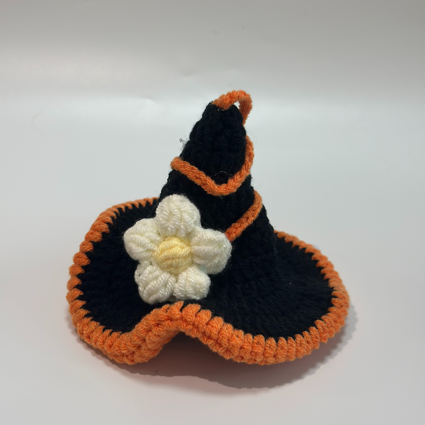 Close-up detail view of handmade crocheted Halloween witch hat for Labubu doll featuring black pointed hat with orange pom-pom, orange scalloped trim edges, and white daisy flower decoration with yellow center. Shows intricate crochet work and floral embellishment quality, perfect Halloween doll hat accessory for collectors and craft enthusiasts.