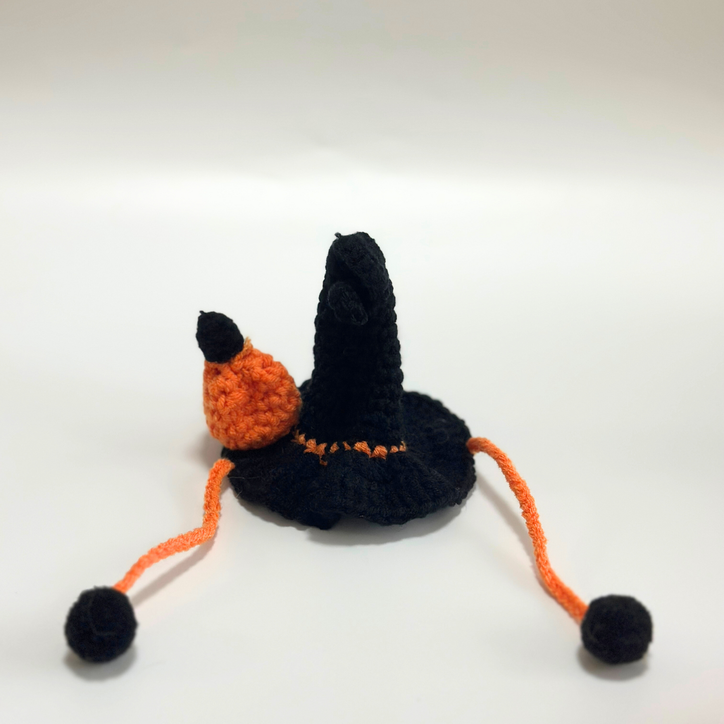 Flat lay view of handmade crocheted Halloween witch hat for Labubu doll costume featuring black pointed hat with orange trim and pom-pom detail, orange drawstring ties with black pom-pom ends. Shows construction details and quality craftsmanship of Halloween doll accessory, perfect collectible item for toy enthusiasts and craft lovers.