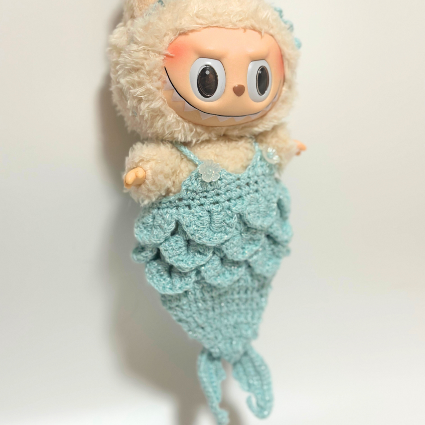 Labubu doll wearing handmade crocheted mermaid tail costume in mint green with textured scale pattern, matching shoulder straps, and decorative shell button detail. Features intricate crochet work creating realistic mermaid tail silhouette with dimensional scales texture, perfect fantasy-themed doll costume for collectors and mermaid enthusiasts.