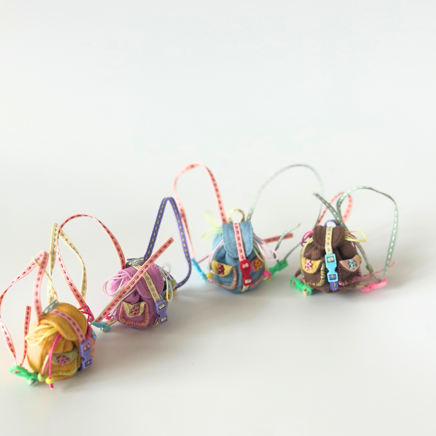 Lineup of handmade miniature backpacks for Labubu dolls in various corduroy colors including yellow, purple, blue, and brown, each featuring unique colorful rainbow straps, metal buckle details, and decorative ribbon accessories. Clean white background showcases the variety and quality craftsmanship of doll backpack collection, perfect for collectors seeking multiple color options.