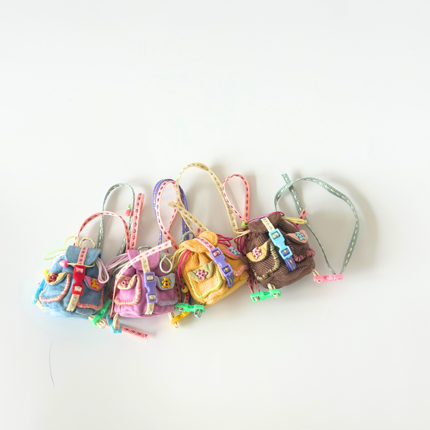 Collection of handmade miniature backpacks for Labubu dolls in various corduroy fabrics including blue, purple, yellow, and brown, each featuring colorful rainbow straps, metal buckle details, front pockets, and decorative ribbon accessories with small charms. Shows variety of color options and intricate craftsmanship of doll backpack accessories, perfect for collectors seeking multiple options.