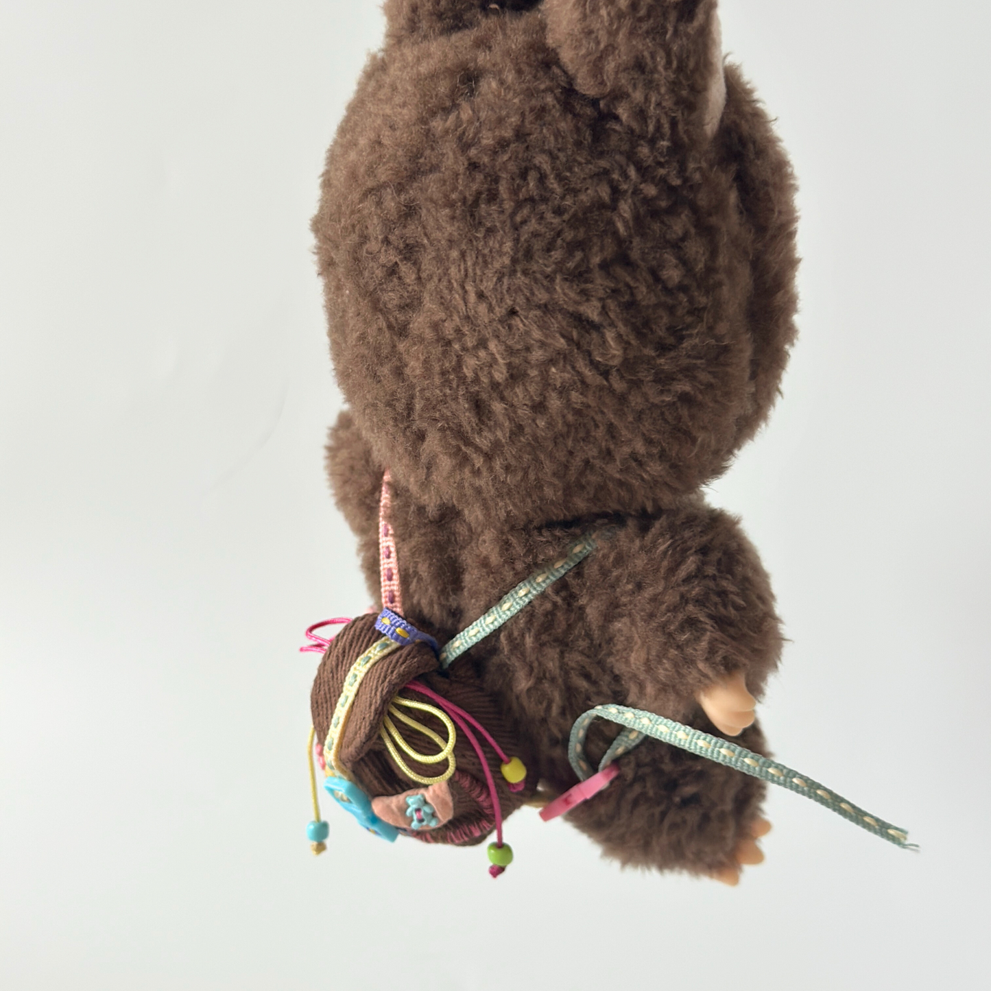 Close-up of brown Labubu doll wearing matching brown corduroy miniature backpack with colorful rainbow straps, decorative ribbons, small charms and beaded accessories. Shows intricate detail of handmade doll accessory construction with functional straps and quality materials, perfect collectible item demonstrating craftsmanship and realistic proportions for toy enthusiasts.