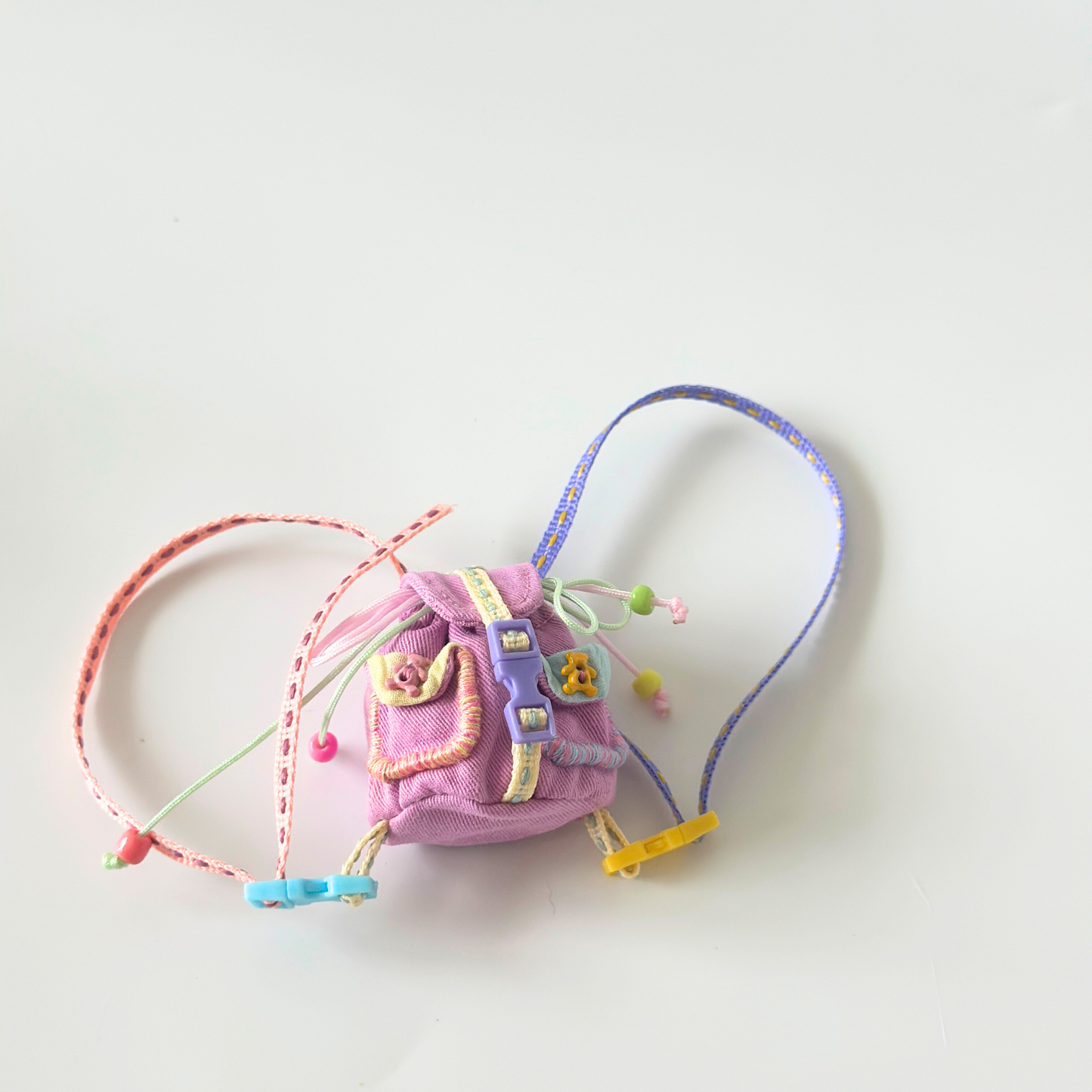Flat lay view of miniature handmade backpack for Labubu doll featuring purple corduroy fabric with front pocket, metal buckle details, colorful rainbow straps in pink, blue, yellow and green with decorative clips, various ribbon accessories and small charms. Shows intricate construction and realistic miniature backpack design, perfect doll accessory for collectors.