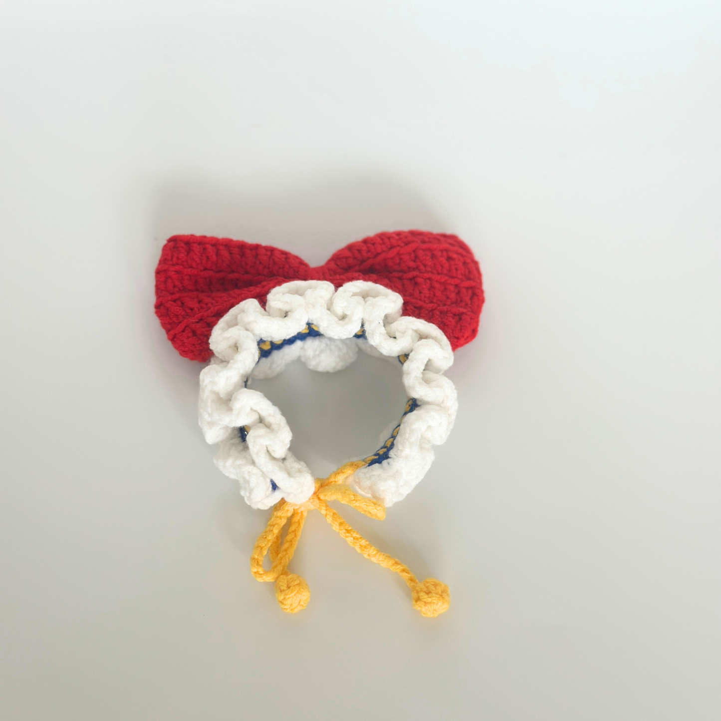 Handmade crocheted Snow White headband for Labubu doll featuring red textured bow, white ruffled trim bonnet with blue accent stitching, and yellow drawstring ties. Perfect doll hair accessory, miniature headpiece, collectible toy fashion item for doll customization and DIY craft projects.