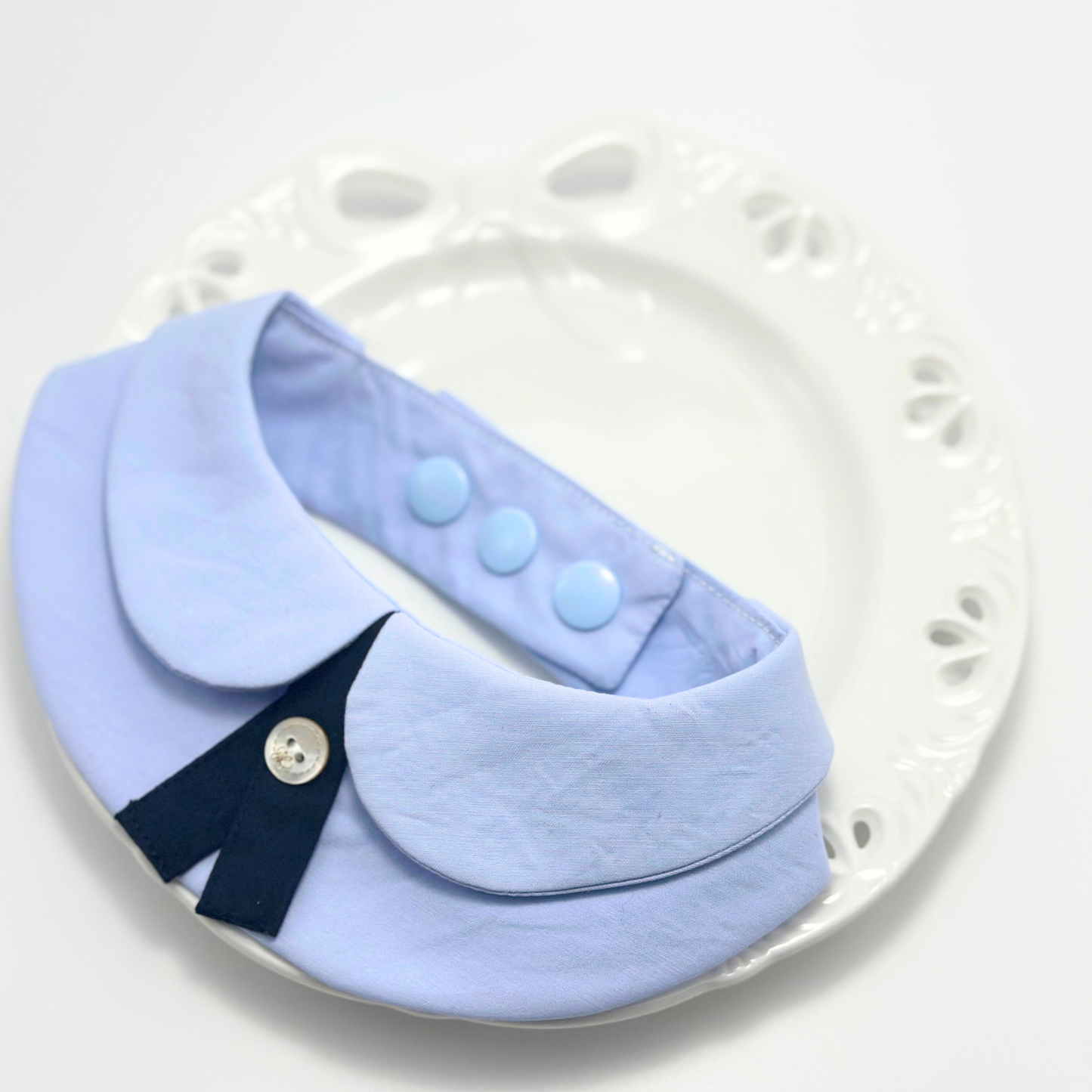 Pet formal collar bandana in blue with snap button adjustable sizing, navy necktie accent displayed on decorative white plate for product photography