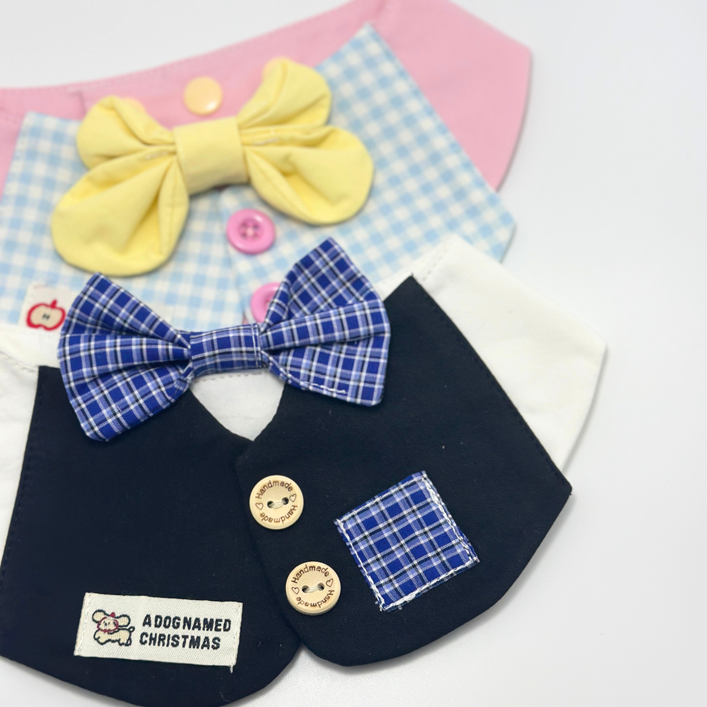 Collection of pet bibs featuring navy blue formal bib with blue plaid bow tie, pink and blue gingham bib with yellow bow, and white bib with cherry pattern - handmade pet accessories with wooden buttons and brand labels