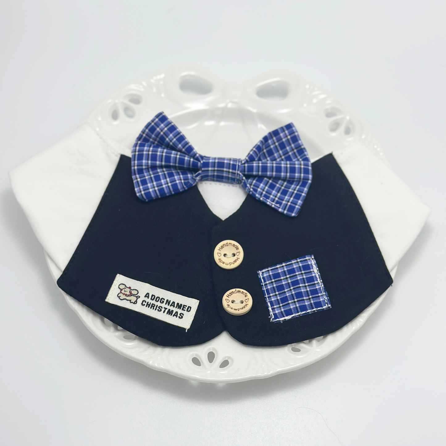 Navy blue pet bib with white collar and blue plaid bow tie displayed on white decorative plate with paw print pattern, featuring "A Dog Named Christmas" label and wooden buttons
