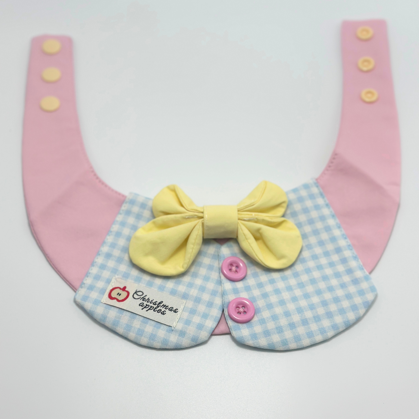 Pink pet bib with blue gingham collar and yellow bow tie laid flat, featuring "Christmas apples" label with red apple logo and pink buttons - cute pet bandana in pastel colors