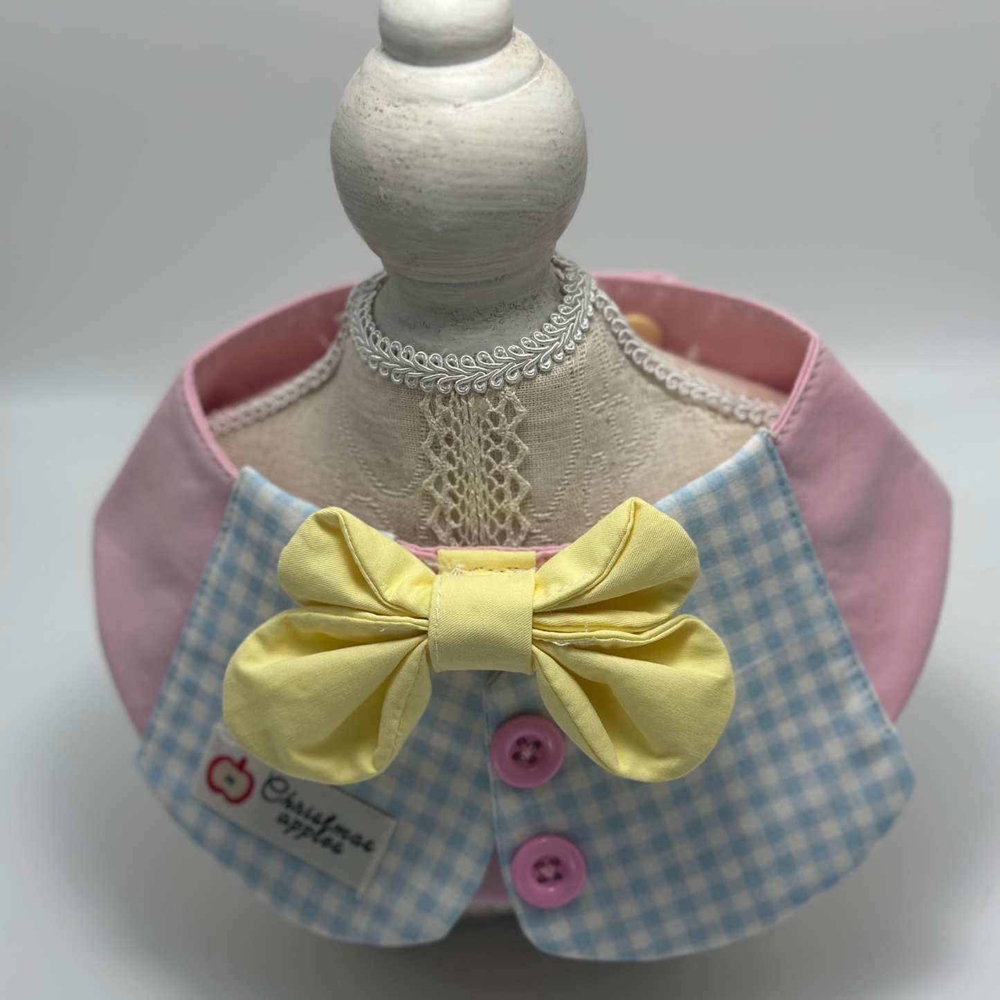 Pink pet bib with blue gingham collar and yellow bow tie displayed on white dress form mannequin with decorative lace necklace, featuring "Christmas apples" label and pink buttons - elegant pet accessory styling