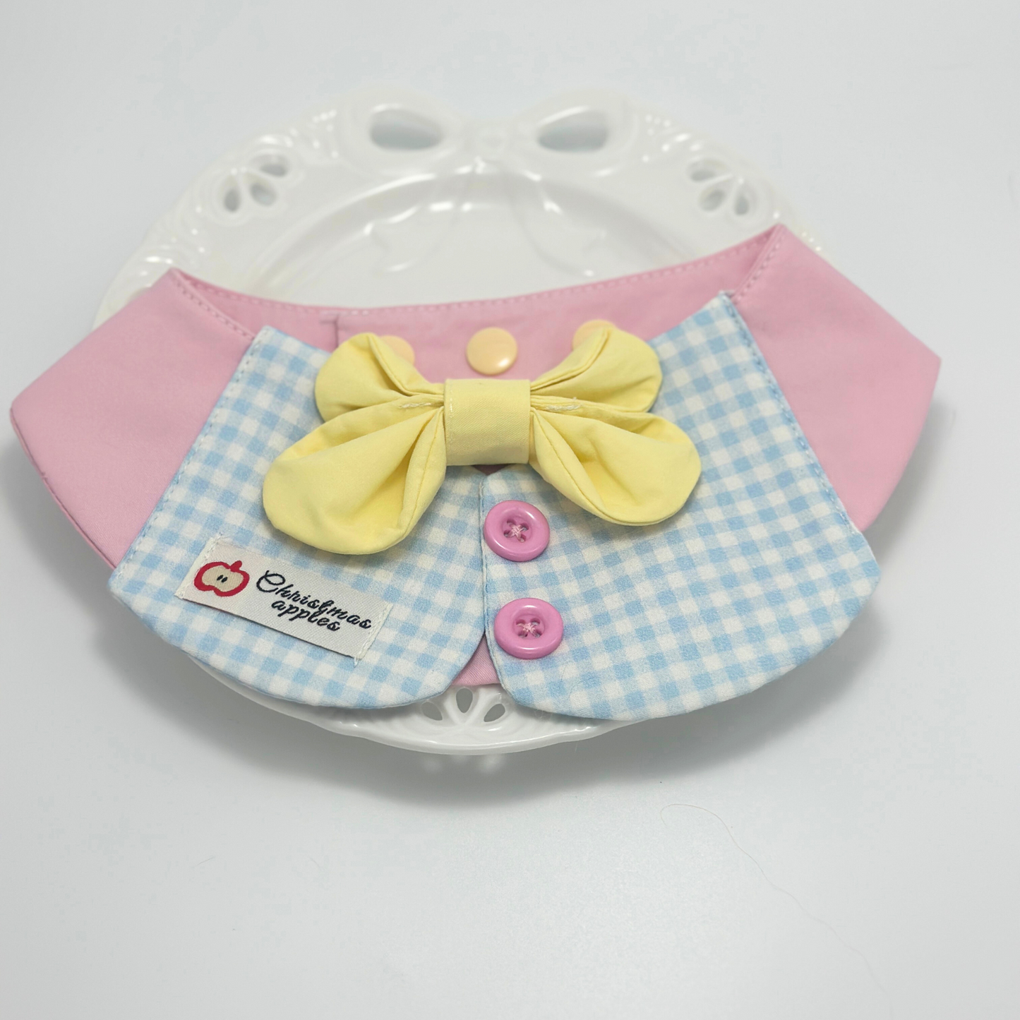 Pink pet bib with blue gingham collar and yellow bow tie displayed on white decorative plate with paw print pattern, featuring "Christmas apples" label and pink buttons - elegant pet accessory presentation