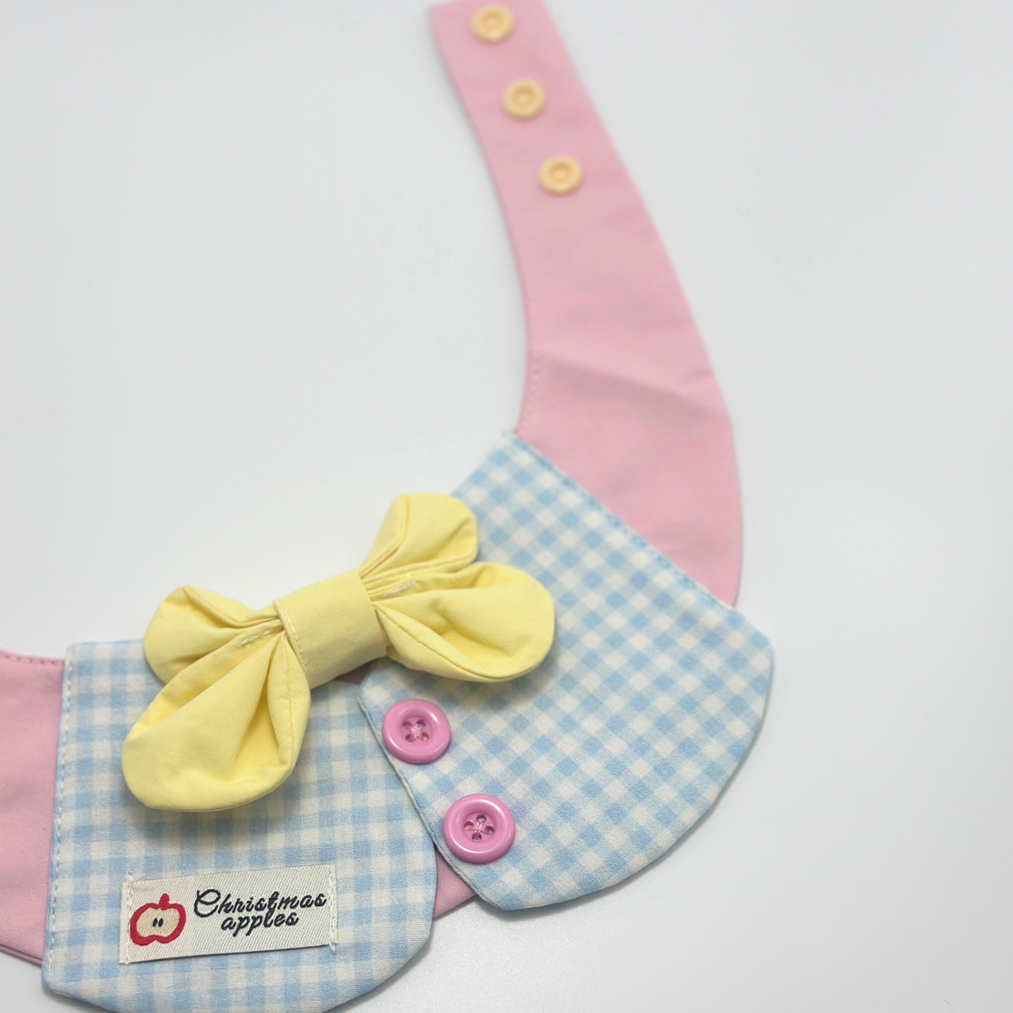 Pink pet bib with blue gingham collar and yellow bow tie on white background, featuring "Christmas apples" label with red apple logo and pink buttons - cute pet bandana product shot