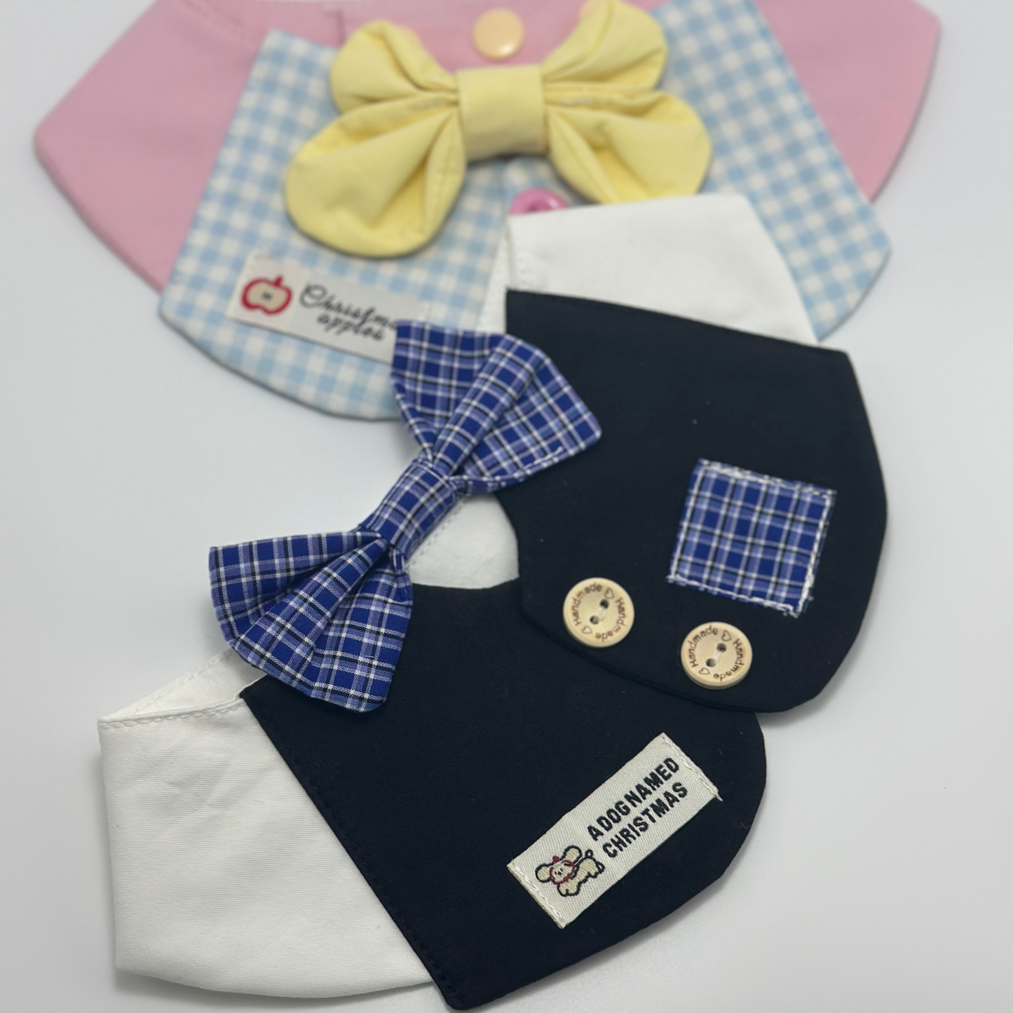 Two pet bibs in overlapping display - navy blue formal bib with blue plaid bow tie and "A Dog Named Christmas" label in front, light blue gingham bib