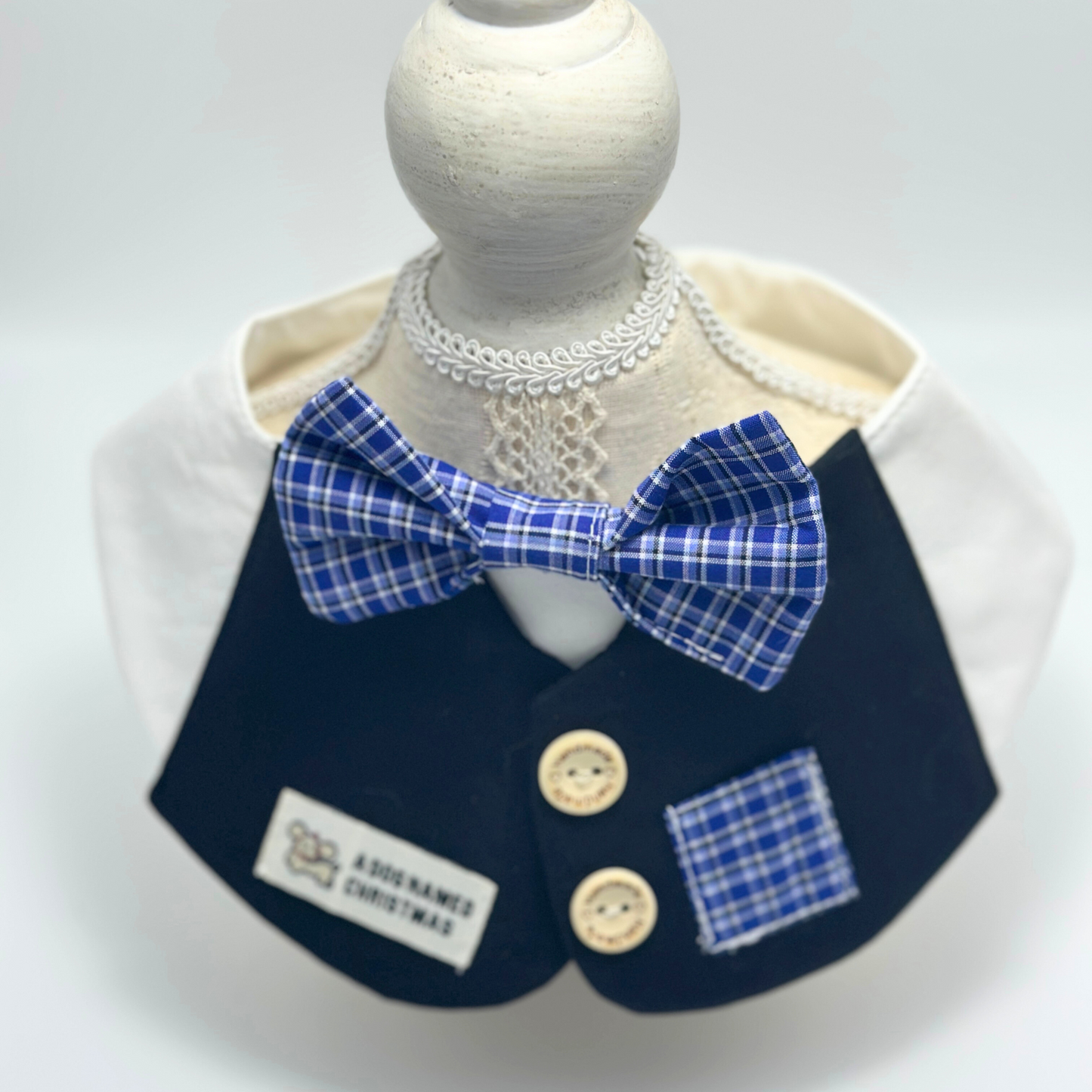 Navy blue formal pet bib with white ruffled collar and blue plaid bow tie displayed on white dress form mannequin with decorative lace necklace, featuring "A Dog Named Christmas" label and wooden buttons