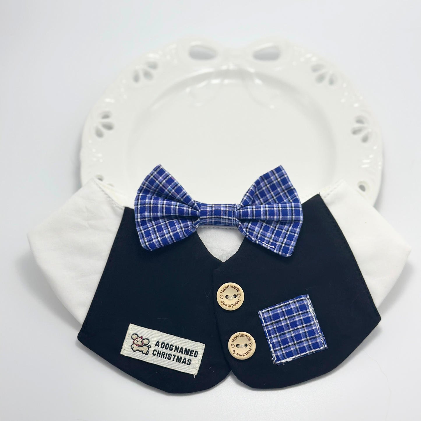 Navy blue formal pet bib with white collar and blue plaid bow tie displayed on white ceramic plate with paw print design, featuring "A Dog Named Christmas" label and handmade wooden buttons