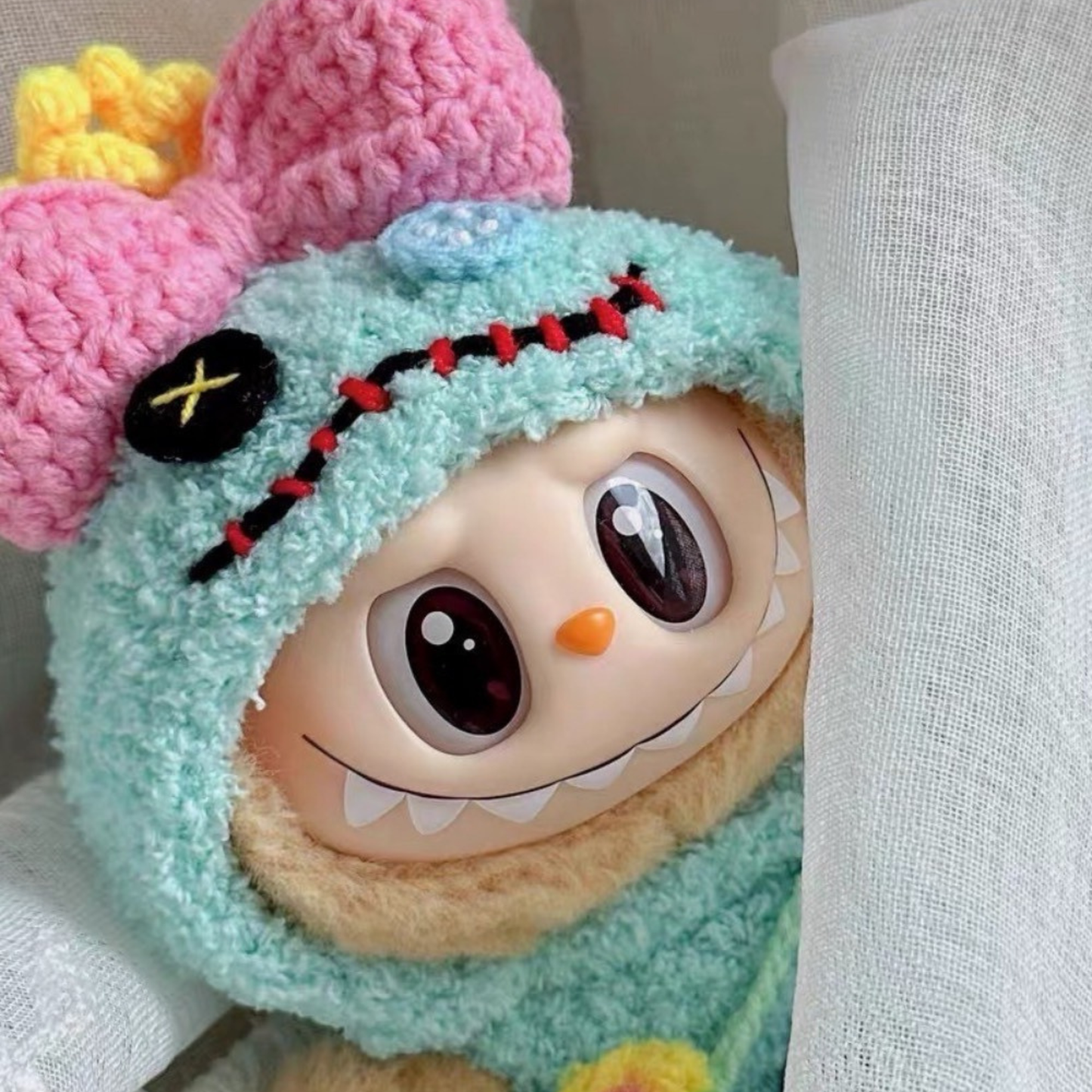 Close-up view of Labubu plush doll wearing handmade teal crochet Scrump costume featuring black button eye patch with yellow X stitching, red and black zigzag smile stitches, and pink bow with yellow crown on top