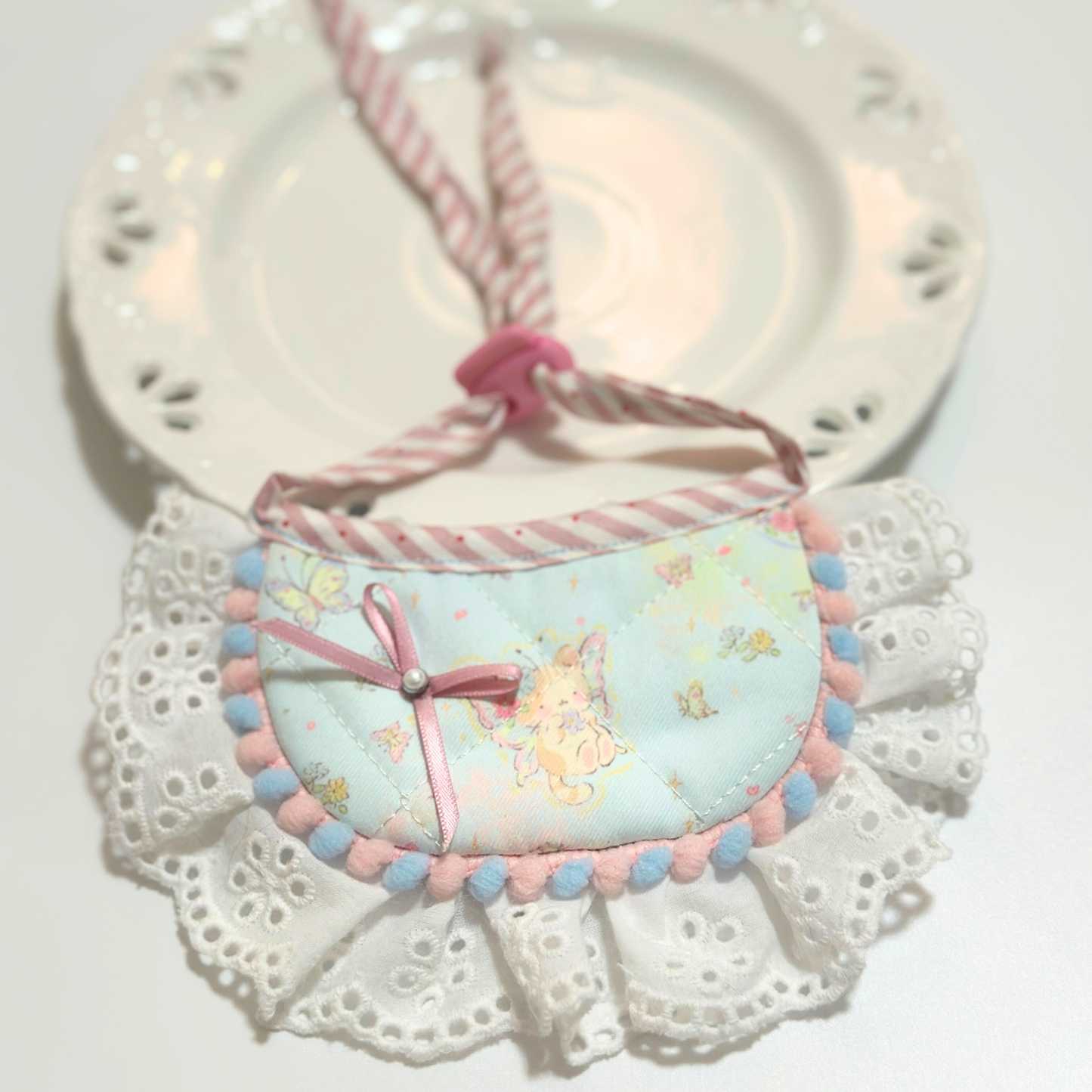Dog bib with pom-pom trim lifestyle photo - Light blue pet bandana bib with bunny pattern, ribbon bow decoration, eyelet lace ruffle, displayed with vintage clock