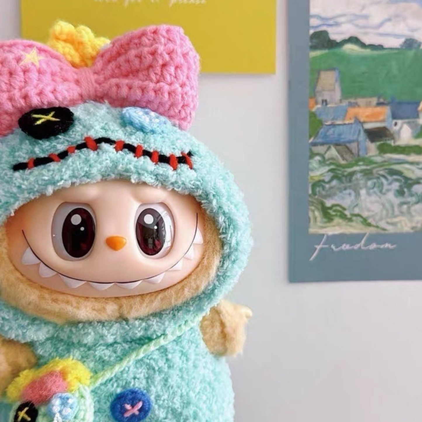 Labubu doll wearing teal crochet Scrump-inspired costume with pink bow and yellow crown, holding miniature Scrump accessory, photographed against a white wall with colorful art prints in background