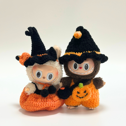 Close-up of two Labubu dolls wearing handmade crocheted Halloween witch costumes with black pointed hats featuring orange pom-poms and trim, orange pumpkin jumpsuits with embroidered jack-o'-lantern faces and green stem details, plus black accessories. Adorable Halloween doll duo, matching seasonal outfits, collectible miniature costumes perfect for Halloween enthusiasts and doll collectors.