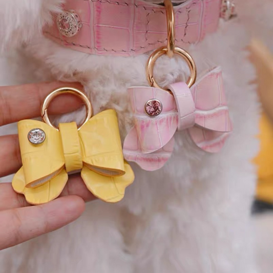 Two decorative pet collar bow accessories with gold keyrings - one in soft yellow and one in pink, both featuring rhinestone embellishments in the center. The bows have a quilted leather texture and are photographed against a fluffy white background with a pink pet collar visible in the scene.