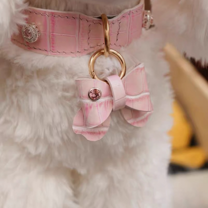 A pink genuine leather bow charm attached to a matching quilted pink pet collar with gold hardware. The bow features a rhinestone center embellishment and is connected via a gold keyring. The collar has decorative stitching and rhinestone studs, photographed against a fluffy white background.
