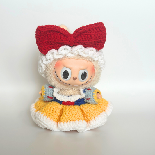 A crocheted Snow White costume for Labubu doll, featuring a white ruffled bonnet with red bow, yellow ruffled skirt, blue bodice with red trim, and white puffy sleeves. The labubu has large round eyes and a sweet expression, displayed against a light gray background