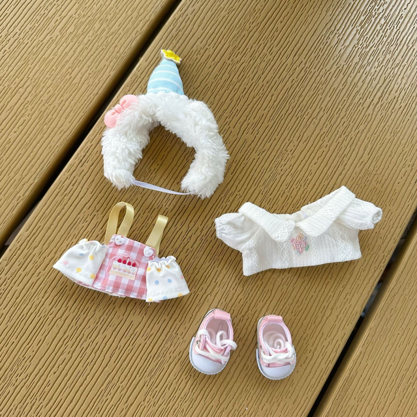 Labubu doll birthday costume accessories laid out separately - fluffy white hat with striped party cap, pink sunglasses, checkered apron with cake print, white ruffled top, and pink sneakers on wooden background