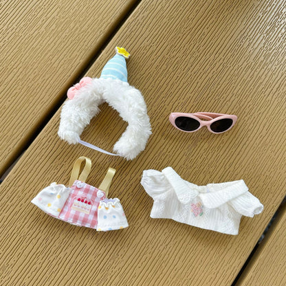 Complete Labubu doll birthday costume accessories laid out separately - white fluffy hood with striped party hat, pink sunglasses, checkered apron with cake print, white ruffled off-shoulder top on wooden deck background
