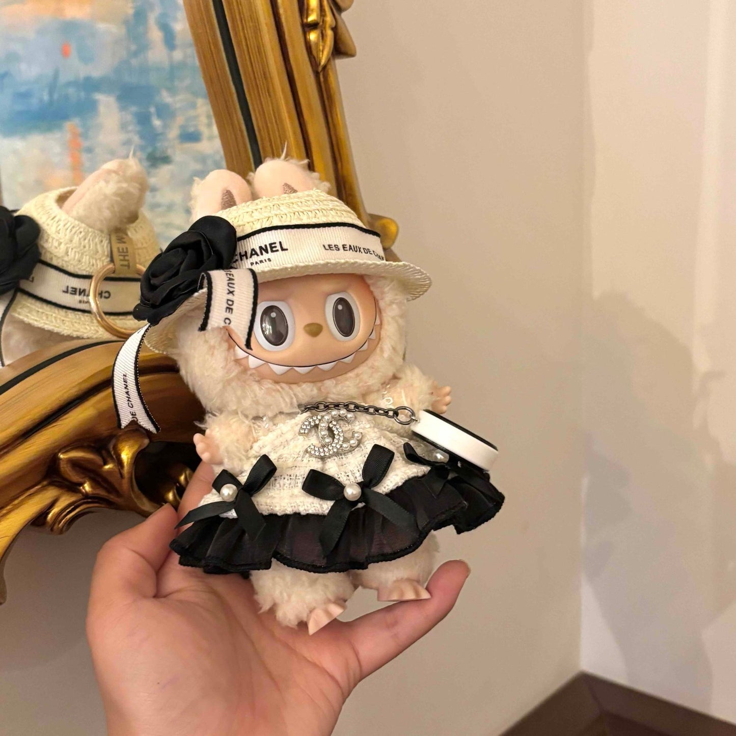 A white Labubu plush doll wearing a miniature Chanel-inspired outfit, including a cream woven hat with Chanel branding and black ribbon, and a black and white ruffled dress with pearl chain details and the iconic Chanel CC logo, held in someone's hand against a mirror background.