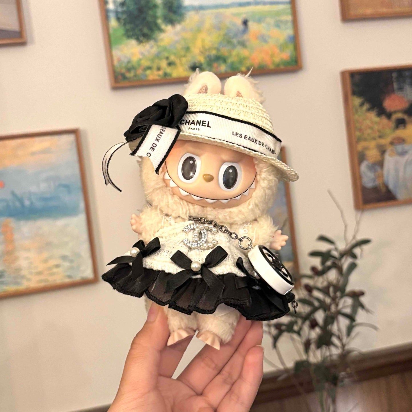 A cream-colored Labubu plush doll wearing a detailed Chanel-inspired outfit including a woven hat with 'CHANEL PARIS' and 'LES EAUX DE CHANEL' ribbons plus black fabric flower, and a white textured dress with rhinestone CC logo, black ruffle hem, and pearl accents with chain details, held in someone's hand against a wall decorated with framed impressionist-style paintings and a small potted plant.