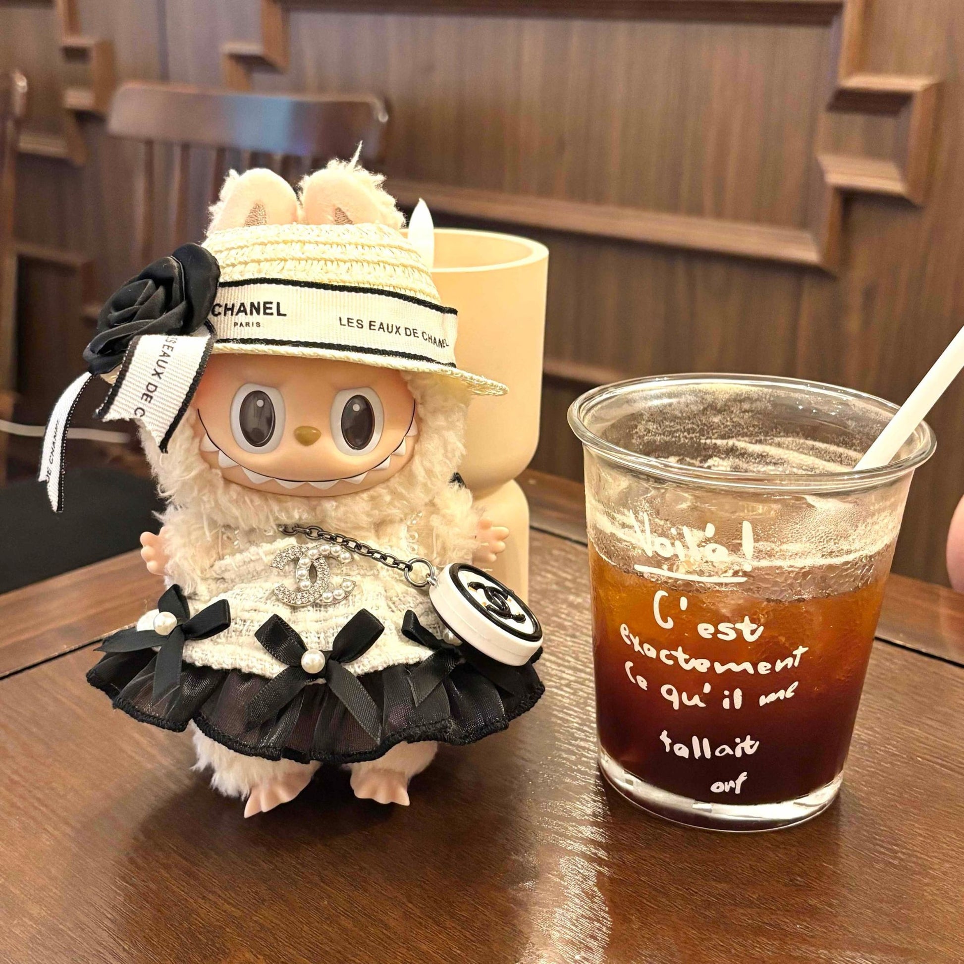 A cream-colored Labubu plush doll in Chanel-inspired outfit sitting on a wooden table next to a glass of iced coffee with French text 'C'est exactement ce qu'il me fallait' (That's exactly what I needed) written on it, with a yellow shopping bag in the background, in a cafe setting with wooden furniture.