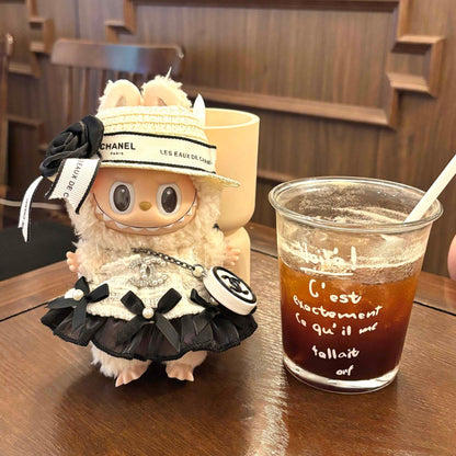 A cream-colored Labubu plush doll in Chanel-inspired outfit sitting on a wooden table next to a glass of iced coffee with French text 'C'est exactement ce qu'il me fallait' (That's exactly what I needed) written on it, with a yellow shopping bag in the background, in a cafe setting with wooden furniture.