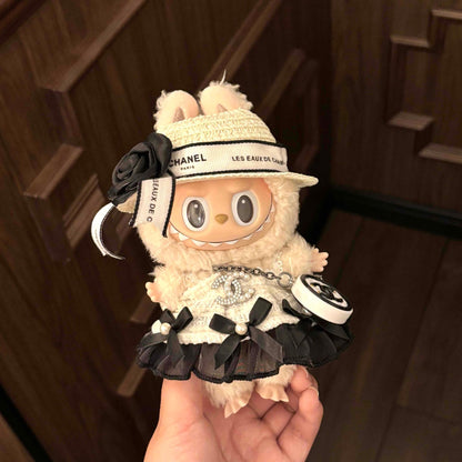 A cream-colored Labubu plush doll in an elaborate Chanel-inspired outfit, featuring a woven hat with 'CHANEL PARIS' and 'LES EAUX DE CHANEL' text bands and black fabric flower, a white textured dress with rhinestone Chanel CC logo, black ruffle trim, pearl details, and a small round Chanel charm, held in someone's hand against a wooden parquet floor background.